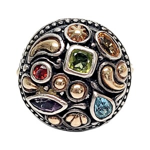 May include: A silver ring with a round, multi-faceted design. The ring is adorned with a variety of colourful gemstones, including green, blue, red, and purple. The ring is also accented with gold details.