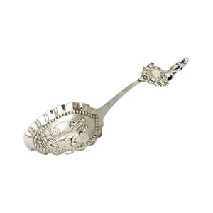 May include: A silver sugar spoon with an ornate handle and a scalloped bowl. The bowl features an embossed design of a woman in a carriage.