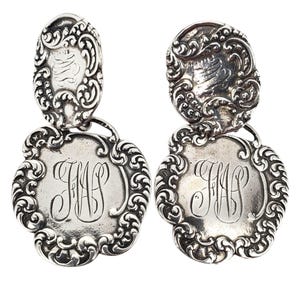 May include: A pair of silver-toned drop earrings. Each earring features an oval top with an engraved design, connected to a larger, floral-edged pendant. The pendants display the engraved initials "JMS".
