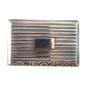 May include: A silver rectangular box with a decorative floral design around the edge. The box has a smooth, flat top with a rectangular, blank area in the center.