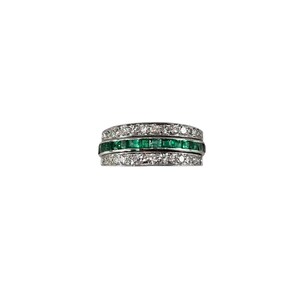 May include: A three-band ring with a central row of emerald-cut green gemstones, flanked by rows of small, clear gemstones. The ring is set in a silver or white metal, creating a striking contrast with the gemstones. The design is elegant and classic.