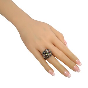 May include: A gold ring featuring a round, multi-coloured gemstone setting. The ring is decorated with intricate details and includes a range of gemstones, such as green, red, blue, and yellow.