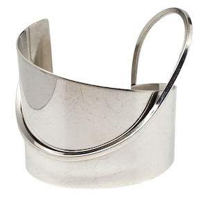 May include: A silver-toned cuff bracelet with a unique design. The bracelet features a wide, curved band with a raised, arched detail on top. The metal has a polished finish, reflecting light.
