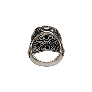 May include: A silver ring with intricate black and gold detailing. The ring has a wide band and a large, ornate top. The inside of the band is marked with the letters "BMS".
