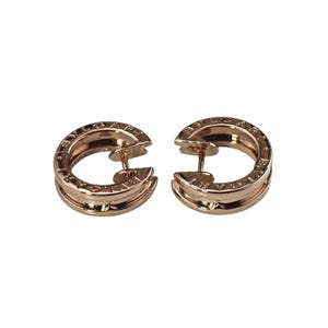May include: A pair of rose gold hoop earrings featuring a circular design with a hollow center. The surface of each earring is engraved with the text BVLGARI along the top and bottom edges, and includes a post-back closure mechanism.
