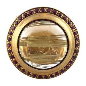 May include: A round gold brooch with a clear, striated stone set in the center. The stone is surrounded by a border of small, dark red gemstones.