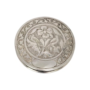 May include: A round, silver-colored compact with an engraved floral and bird design. The central motif features a flower and two birds, surrounded by ornate scrollwork. The compact has a hinged lid and a smooth, reflective surface.