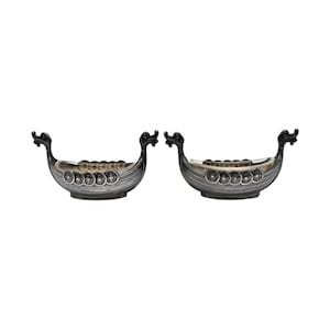 May include: Two silver-toned Viking ship-shaped decorative bowls. Each bowl features a detailed design with a dragon head at the bow and stern. The bowls have a series of circular embellishments along the sides, adding to their historical aesthetic.