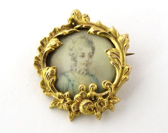 May include: A gold-toned brooch with an ornate, floral design. A miniature portrait of a person is visible in the center of the brooch.