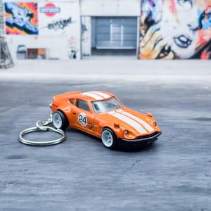 May include: Orange and white toy car keychain. The miniature car features a classic sports car design with white racing stripes and the number 24. The keychain has a silver ring.