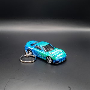 May include: A blue and green toy car keychain with white lettering. The car is a Mazda RX-7 and has a silver key ring.