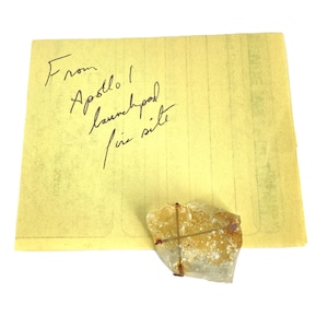 May include: A piece of rock with a wire cross, attached to a yellow sticky note. The note has handwritten text that reads "From Apollo 1 launchpad fire site."