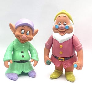 May include: Two plastic figurines of cartoon characters. One is wearing a green outfit with a purple hat and shoes. The other is wearing a pink outfit with a yellow hat and glasses. Both have smiling faces.