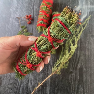 May include: Three bundles of dried green cedar smudge sticks tied with red string. The smudge sticks are on a dark wood surface. One smudge stick is lit and smoking.