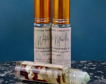 May include: Two gold-capped glass bottles with a white label that reads "Living Roots Dots Creations Water Maiden A Rusalka-Inspired Oil for Empowerment & Confidence 10ml / .33oz Ingredients: Herbal Infused Sweet Almond Oil Essential Oils of Sandalwood, Jasmine"