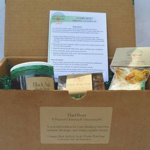 Könnte beinhalten: Ein brauner Karton mit einem grün-weißen Etikett, auf dem "Hard Reset: A Personal Cleansing & Unlocking Kit" steht. Die Box enthält ein Glas mit schwarzem Salz-Körperpeeling, eine Seife und eine Tüte mit Badesalz.