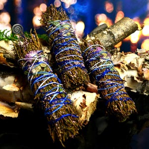 May include: Three bundles of dried herbs and twigs wrapped in vibrant blue and white string. Each bundle features small purple flowers and green foliage, secured tightly with the colorful thread, creating a textured, cylindrical arrangement.