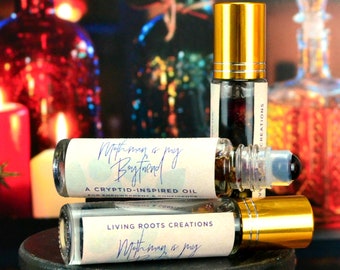 May include: Three glass bottles with gold lids and labels that read "Mothman is my Boyfriend" and "Living Roots Creations". The bottles are filled with a clear liquid and contain small, dried flowers. The bottles are sitting on a black surface.