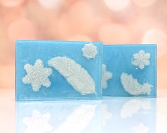 May include: Two rectangular bars of blue soap with white snowflake and feather designs. The soap has a translucent appearance, with the designs slightly raised on the surface. The background is a soft, out-of-focus bokeh.
