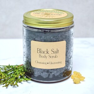 以下が含まれることがあります： 黒い塩のボディスクラブが入ったガラス瓶。瓶には金色の蓋と、"Black Salt Body Scrub Cleansing Uncrossing"と書かれた茶色のラベルが付いています。