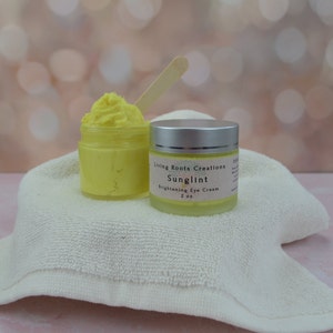Puede incluir: Un pequeño tarro de crema para ojos amarilla con la etiqueta "Living Roots Creations Sunglint Brightening Eye Cream 2 oz."