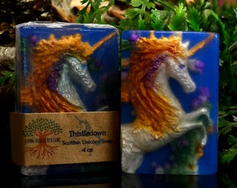 May include: Two rectangular bars of blue soap featuring a raised, three-dimensional white unicorn with a golden mane and horn. A brown paper label wrapped around one bar displays the text Living Roots Creations, Thistledown Scottish Unicorn Soap, 4 oz.