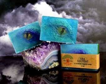 May include: Three bars of handmade soap with a sea serpent design. The soap is blue and green with a gold eye. The soap is on a purple and white crystal. The label on the soap reads "Sea Serpent, a mystical organic soap, 4 oz".