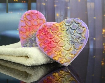 May include: Two heart-shaped bath bombs with a mermaid scale design in shades of pink, purple, and gold. The bath bombs are sitting on a white towel.