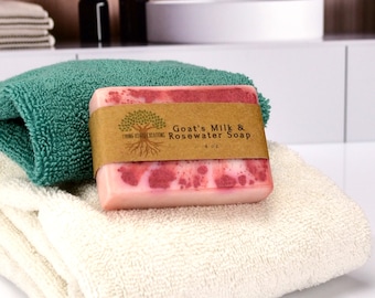 May include: A pink and white bar of soap with a brown label that reads "Goat's Milk & Rosewater Soap" and "Living Essential Creations" on a white towel with a green towel underneath.