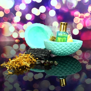 Puede incluir: Un cuenco azul turquesa con una superficie texturizada contiene una pequeña botella de líquido verde con una tapa dorada. El cuenco está sobre una superficie negra con flores secas, un cristal negro y piedras preciosas rojas.