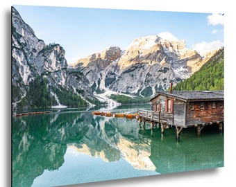 Può includere: Stampa di un paesaggio con una capanna di legno su palafitte su un lago. Il lago riflette le montagne e il cielo. Le montagne sono innevate e l'acqua è di un limpido colore turchese.