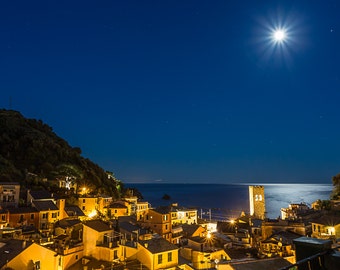 Può includere: Vista notturna di un piccolo paese su una collina che domina l'oceano. Il paese è illuminato da luci, e la luna splende brillantemente nel cielo.