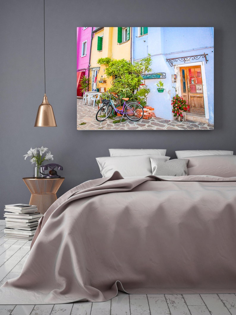 Pu&ograve; includere: Una stretta via colorata in una citt&agrave; europea con una bicicletta parcheggiata in mezzo alla strada. Gli edifici sono dipinti in tonalit&agrave; di rosa, giallo, blu e verde. Ci sono piante in vaso e fiori davanti agli edifici.