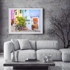 Pu&ograve; includere: Un divano bianco con due cuscini si trova di fronte a un muro grigio con una stampa incorniciata di un edificio colorato con una bicicletta di fronte. Un albero spoglio &egrave; sullo sfondo.