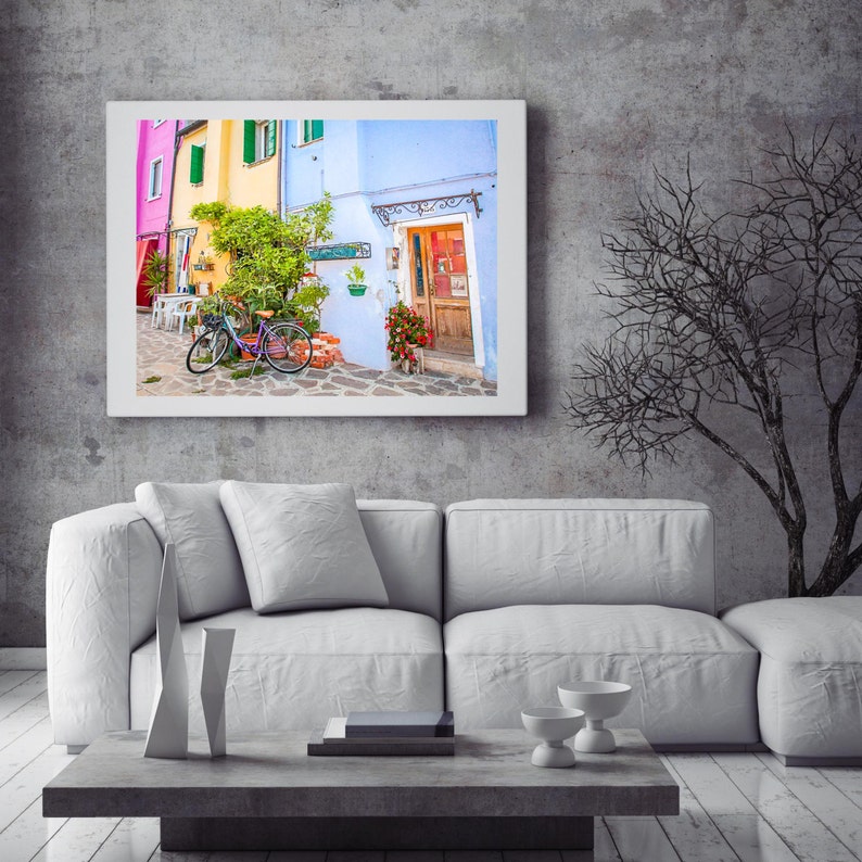 Pu&ograve; includere: Un divano bianco con due cuscini si trova di fronte a un muro grigio con una stampa incorniciata di un edificio colorato con una bicicletta di fronte. Un albero spoglio &egrave; sullo sfondo.