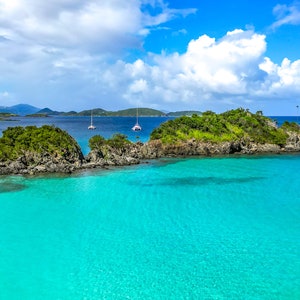 Op de afbeelding: Een panoramisch uitzicht op een tropisch eiland met turquoise water, weelderige groene vegetatie en witte zandstranden. Zeilenboten liggen voor anker in het heldere water.