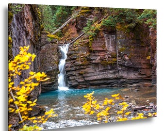 Puede incluir: Fotografía de un paisaje de una cascada que cae en una piscina turquesa. La cascada está rodeada de acantilados de roca roja en capas y árboles verdes. El follaje amarillo enmarca el primer plano.