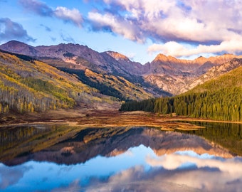May include: A mountain lake with a reflection of the surrounding mountains and clouds in the water. The mountains are covered in trees, some of which are changing color for fall.