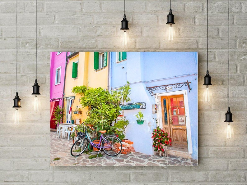 Pu&ograve; includere: Un edificio colorato in stile vintage con una porta blu e una bicicletta parcheggiata davanti. L'edificio ha una facciata rosa, gialla e blu con persiane verdi. La bicicletta &egrave; viola e ha un cestino sul manubrio.