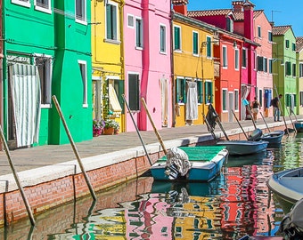 Può includere: Una vivace scena di strada con edifici colorati in tonalità di verde, giallo, rosa, arancione e rosso. Le barche sono ormeggiate lungo un canale, con riflessi degli edifici nell'acqua. Il cielo è azzurro.