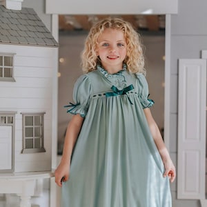 Puede incluir: Un camisón azul claro con mangas cortas, dobladillo con volantes y ribete verde oscuro. El camisón presenta un cuello alto con un lazo verde oscuro y detalles de encaje. La niña lleva el camisón y sonríe.
