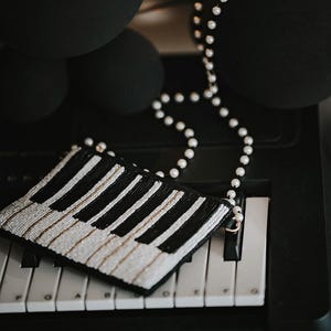 May include: A black and white beaded clutch resembling piano keys rests on a keyboard. A pearl necklace and black spheres are in the background. The clutch has a gold zipper and is a unique accessory.