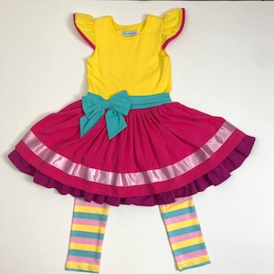 Pu&ograve; includere: Un vestito giallo senza maniche con un fiocco turchese e una gonna rosa con un nastro di raso rosa. L'abito &egrave; abbinato a leggings a righe in giallo, rosa e turchese.