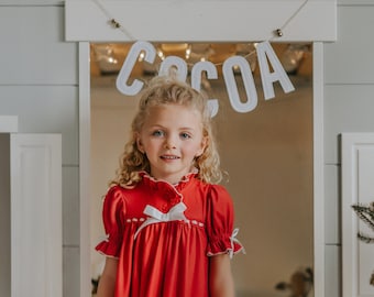 Könnte beinhalten: Ein junges Mädchen trägt ein rotes Nachthemd mit weißer Bordüre und einer weißen Schleife. Das Nachthemd hat kurze Ärmel und einen Rüschensaum. Das Wort "COCOA" ist in weißen Buchstaben über dem Mädchen geschrieben. Das Mädchen steht in einer Tür.