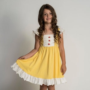 May include: A young girl wearing a yellow dress with white ruffles and red buttons. The dress has a white bodice with a yellow skirt. The girl has long brown hair and is smiling.