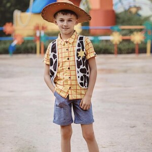 Puede incluir: Un ni&ntilde;o peque&ntilde;o vestido como Woody de Toy Story. Lleva una camisa de cuadros amarillos y rojos, un sombrero de vaquero marr&oacute;n, un chaleco marr&oacute;n y blanco, pantalones cortos de mezclilla azul y zapatillas negras. Est&aacute; de pie frente a un &aacute;rea de juegos colorida.