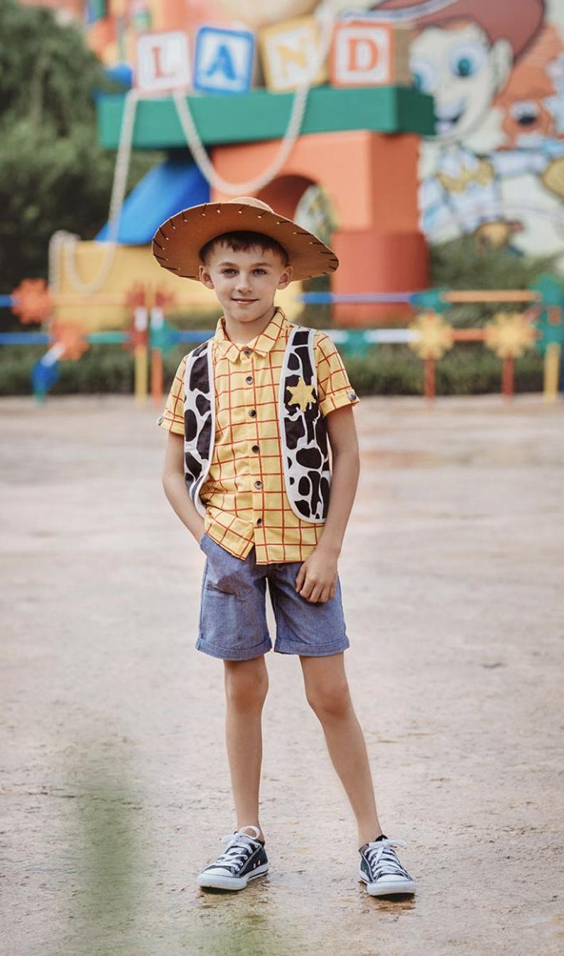 Puede incluir: Un ni&ntilde;o peque&ntilde;o vestido como Woody de Toy Story. Lleva una camisa de cuadros amarillos y rojos, un sombrero de vaquero marr&oacute;n, un chaleco marr&oacute;n y blanco, pantalones cortos de mezclilla azul y zapatillas negras. Est&aacute; de pie frente a un &aacute;rea de juegos colorida.