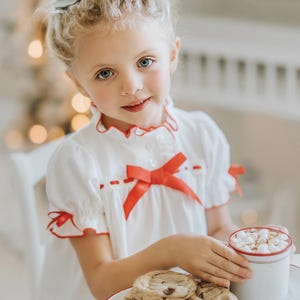A young girl is wearing The Original Clara Nutcracker Christmas Gown Short Sleeve in White, featuring red accents, a bow, and a ruffled hem from the Christmas Lounge Collection. The buttery soft fabric is CPSC certified.