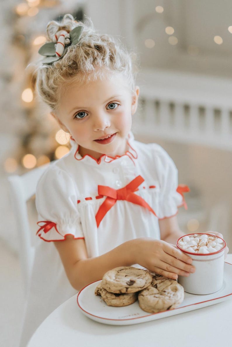 A young girl is wearing The Original Clara Nutcracker Christmas Gown Short Sleeve in White, featuring red accents, a bow, and a ruffled hem from the Christmas Lounge Collection. The buttery soft fabric is CPSC certified.