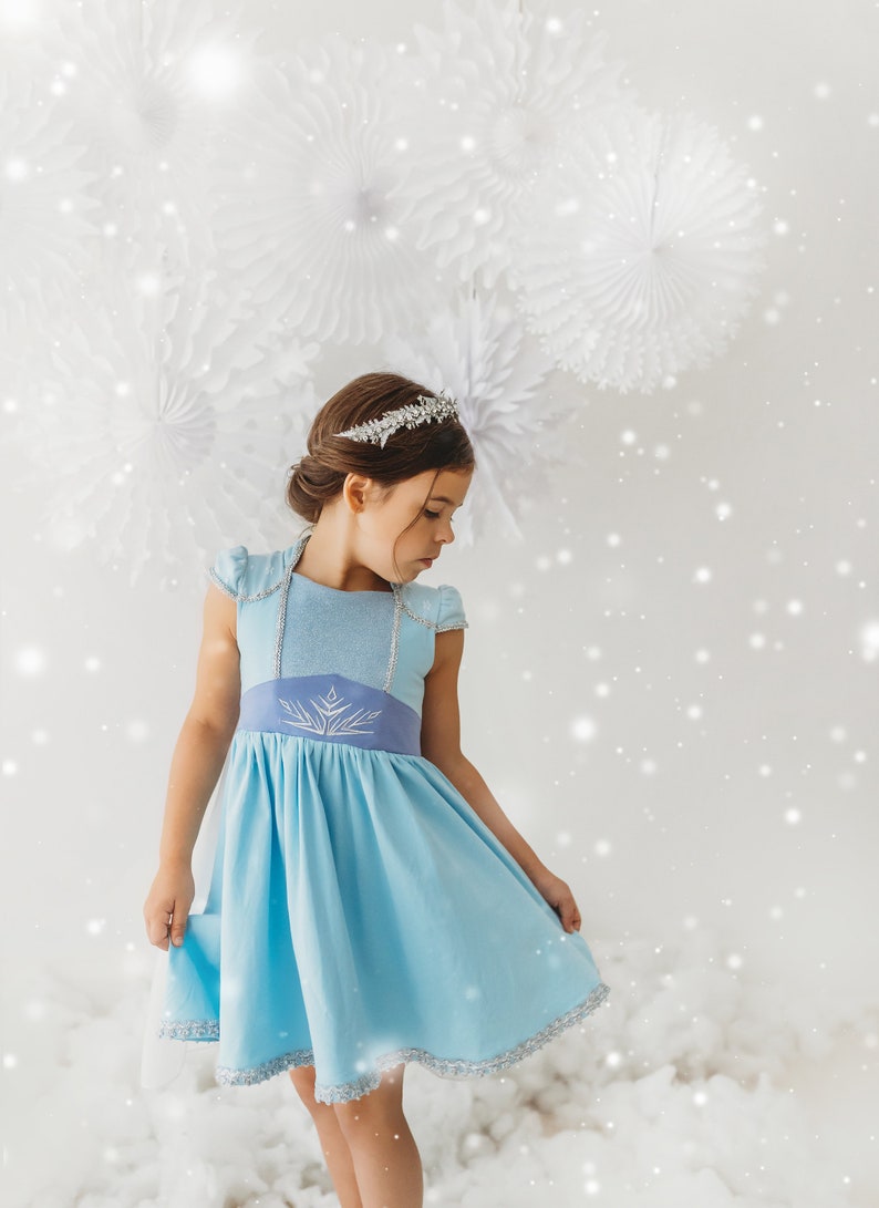 May include: A light blue dress with a silver trim and a silver tiara. The dress has a light blue bodice and a darker blue waistline with a silver snowflake design. The dress is a costume, likely for a child.